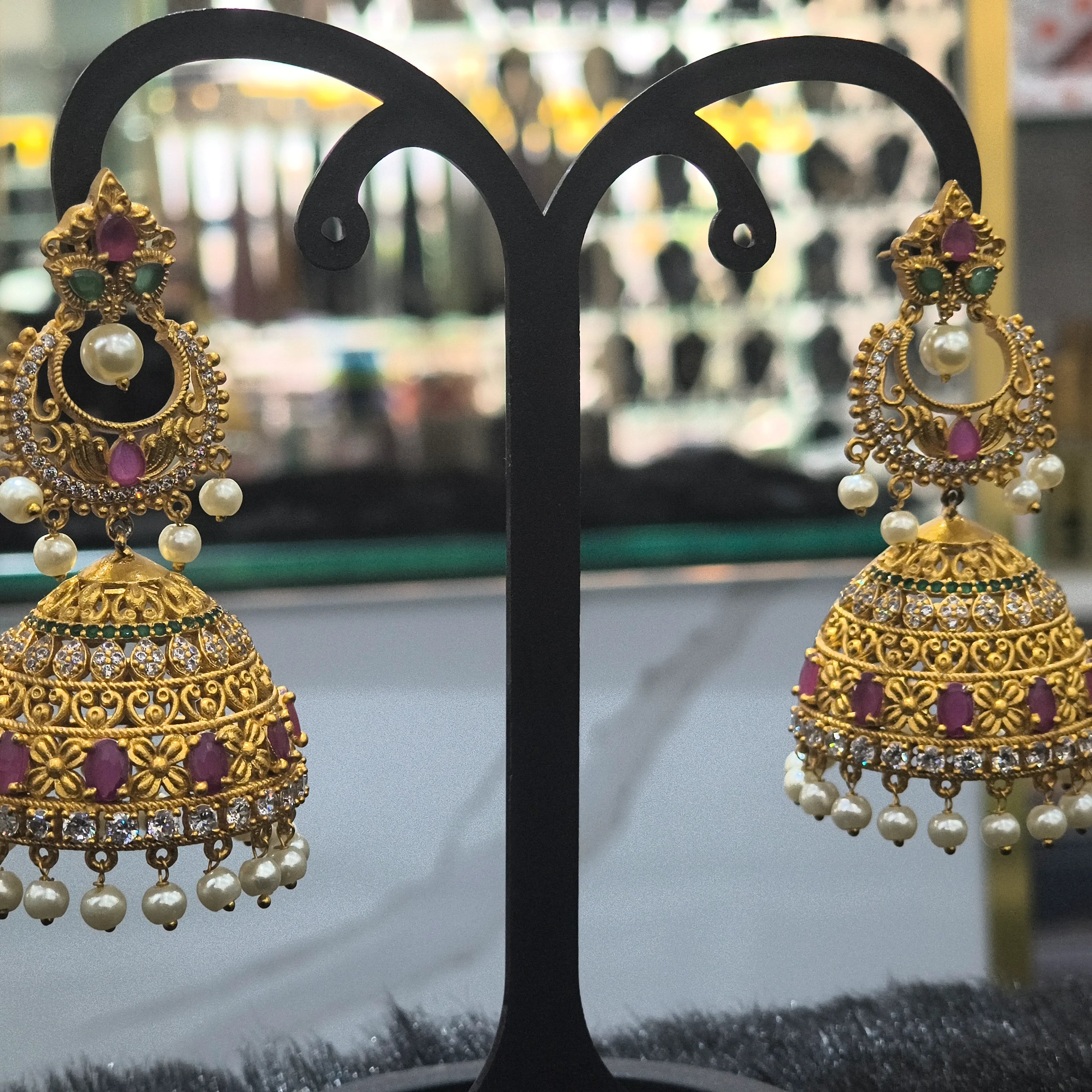 Ornate gold earrings with pearls and gemstones on a black stand.