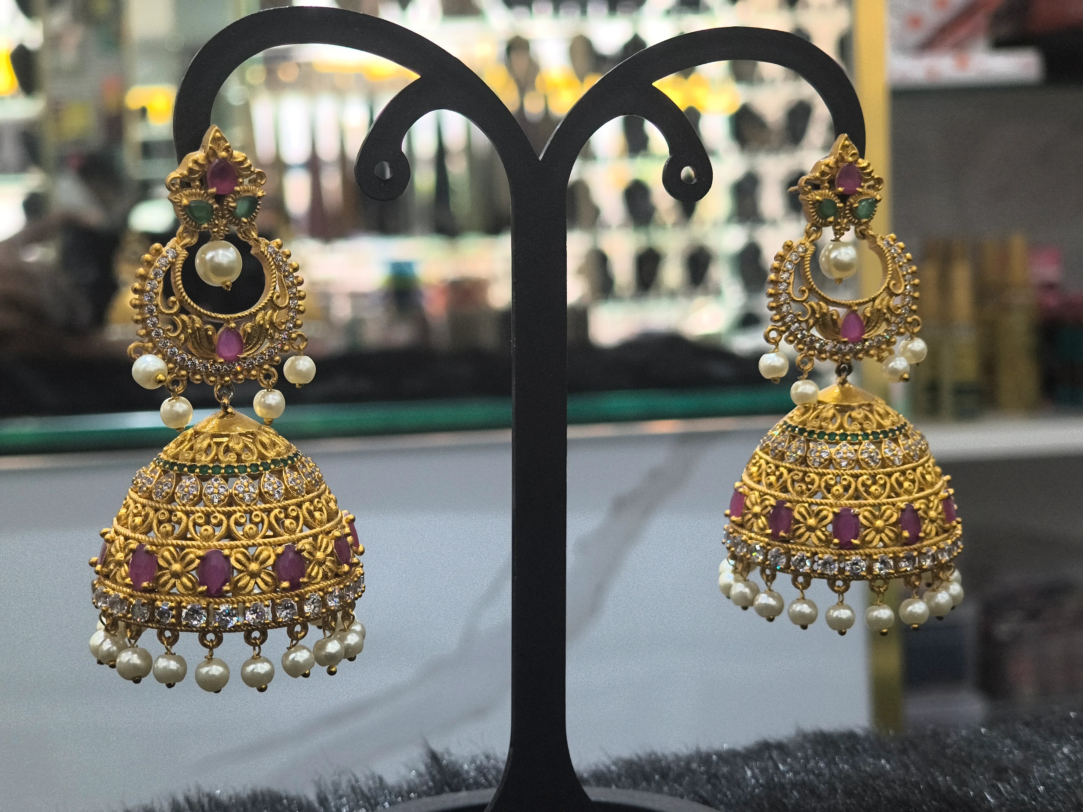 Ornate gold earrings with pearls and gemstones on a black stand.
